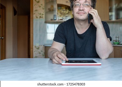 A Man In The Glasses Is Sitting At A Table At Home. Office At Home. The Man Is Talking On The Phone And At The Same Time Smiling. Not A Model, Just An Ordinary Person. Man Looking Far