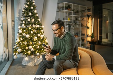 Man with glasses smiling while checking his mobile phone, sitting on a modern yellow sofa in a festive decorated office with a christmas tree during the holiday season