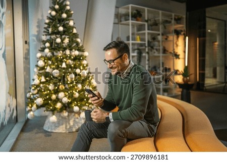 Man with glasses smiling while checking his mobile phone, sitting on a modern yellow sofa in a festive decorated office with a christmas tree during the holiday season