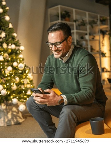 Man with glasses smiling while checking his mobile phone, sitting on a modern yellow sofa in a festive decorated office with a christmas tree during the holiday season