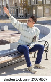 Man With Glasses In White Sweater Siting On Bench And Holding Newspaper  Next To Fountain In Town And Waving Someone Near