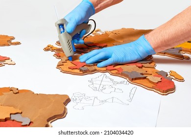 A Man Glues A Wooden Model Of The Country To The World Map In Russian Using A Glue Gun.