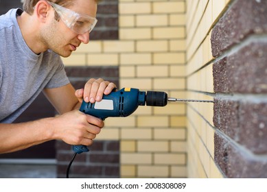 A man in goggles drills a brick wall with a drill. Do it yourself