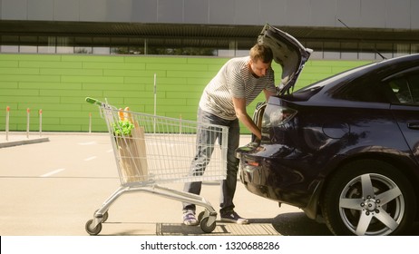 Man Going With Cart On Shopping Mall Parking. Young Caucasian Smiling Male Putting Paper Bags With Food And Watermelon Into Car Trunk.