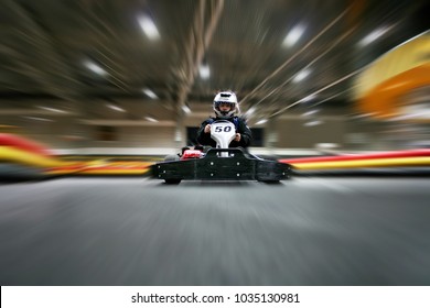The man is going on the go-kart on karting track indoors. He is wearing a helmet.