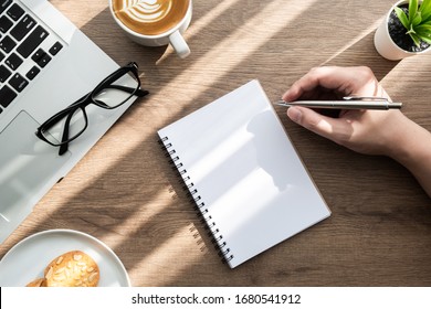 Man is going to write something on blank notebook page on wood office desk table with cookies, cup of coffee and supplies. Morning life at work concept, top view.