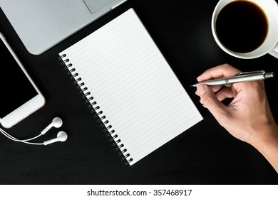 A man is going to write something on a notebook. Black office table with smart phone, laptop, a cup of coffee and supplies. Top view.