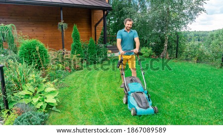 A man in a good mood with a smile mows the gas using an electric lawn mower. Beautiful lawn in the landscape design of a country house. Seasonal garden care.