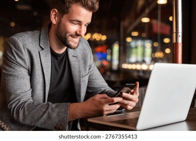 Man in a gray blazer using a smartphone while seated at a laptop in a cozy cafe with warm lighting and blurred background.