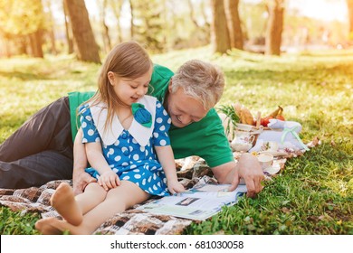 Man With Gray Hair Laying On Blanket Holding His Granddaughter Reading Magazine On Picnic On Summer. 