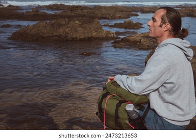 A man in a gray hoodie meditates on a rocky beach by the ocean, embracing the serene environment. He holds a green backpack, suggesting an adventure or hiking trip, amid the tranquil coastal setting. - Powered by Shutterstock