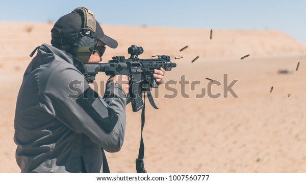 Man with gray jacket shooting black rifle\
on range in desert shells ejecting\
angled