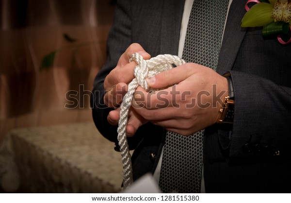 A man in a gray suit and tie tries\
to untie a hard knot on a white cord  with his\
hands.
