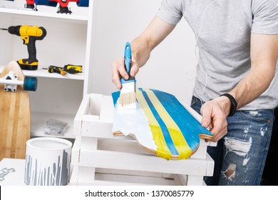 A man in a gray T-shirt is painting a skateboard board in a workshop and listening to music on headphones. - Powered by Shutterstock