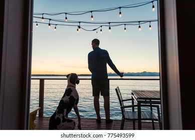 Man And Great Dane Dog Enjoying A Sunset Over Sea View With A Glass Of Wine, Rear View From Window.