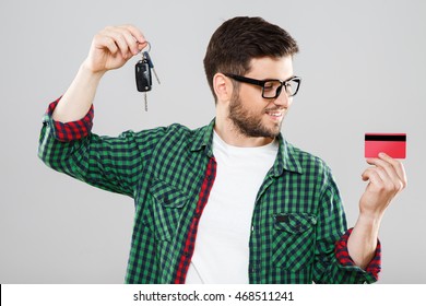 Man In Green Checked T-shirt And Eyeglasses Holding Car Keys In One Hand And Red Credit Card In Other. Looking At Credit Card. Waist Up. Indoors, Studio