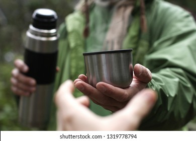 man in green jacket in nature offers a hot drink - tea or coffee from thermos to someone who pulls a hand, first person view, stay for rest in hike or travel   - Powered by Shutterstock