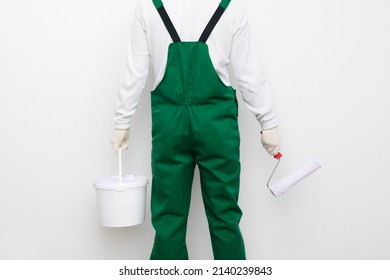 A Man In A Green Overalls Holds A Bucket Of Paint And A Brush In His Hands Against A White Wall, Rear View