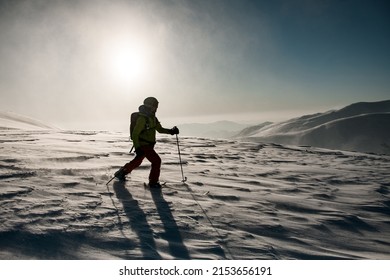 man in green and red ski suit with trekking poles walks through deep powdery snow on a mountain slope.Bright sun lights the snow-capped mountains in the background