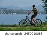 A man in a green T-shirt and black shorts with a backpack on a gray electric mountain bike in motion in the background Lake Garda, mountain peaks in the snow.