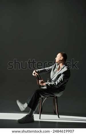 A man in a grey suit sits on a chair, bathed in light.
