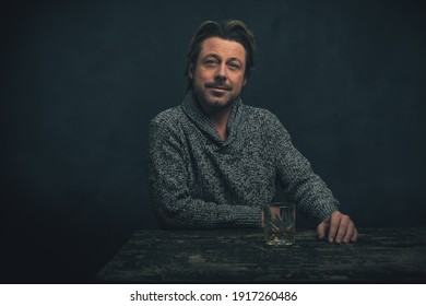 Man In Grey Woolen Sweater With Glass Of Whiskey Behind Old Worn Out Wooden Table.