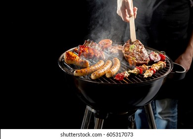 Man Grilling An Assortment Of Meat And Kebabs On A Portable Barbecue Lifting A T-bone Steak With A Pair Of Tongs In A Close Up View Of His Hand