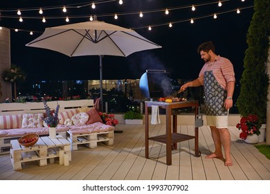 A Man Grilling Barbecue On Rooftop Patio, Enjoys Warm Summer Night At Home Courtyard