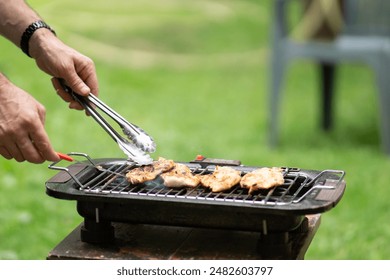 Man grilling flipping meat on barbecue grill grid in his backyard. close up of man making barbecue in his backyard, a man uses tongs to flip a grilled meat on a barbecue grill, green grass in the back