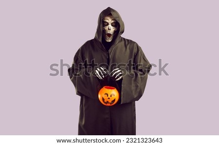 Man in grim reaper costume with creepy facial expression collects candy during Halloween celebration. Man in black robe and skeleton makeup holds candy bucket in form of pumpkin on lilac background.
