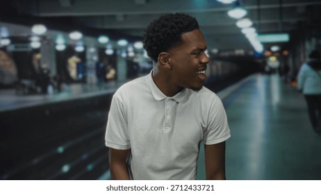 Man grimaces on empty train station platform wearing white polo shirt; frustration delay commute hurry.