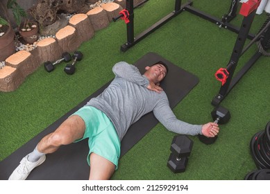 A man grimaces in pain after suffering a minor pectoral tear or chronic rotator cuff injury. Lying on a black mat at a home gym. Workout injury.
