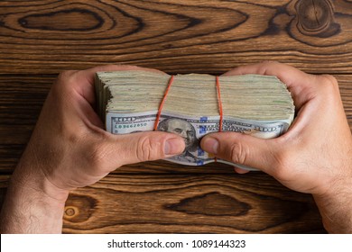 Man Gripping A Thick Stack Of 100 Dollar Bills Or Benjies Tightly Between His Fingers Over A Wooden Table In A Conceptual Overhead Image