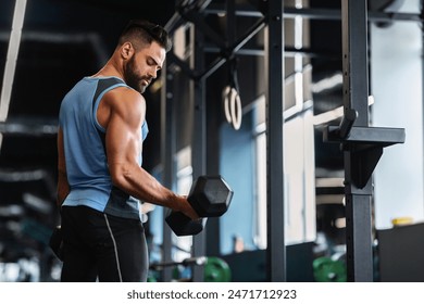 A man in a gym setting lifts a dumbbell during a workout. He is wearing a sleeveless shirt and black pants. - Powered by Shutterstock