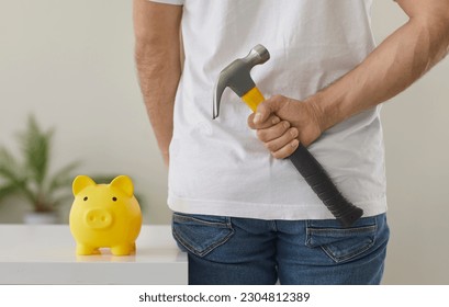 Man with hammer wants to break piggy bank and take out his money savings. Crop backside view man going to smash yellow piggy bank on table with tool of destruction that he is holding behind his back