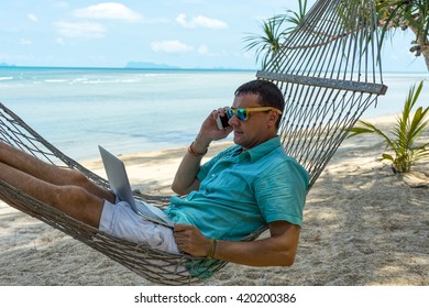 Man In A Hammock On A Tropical Beach Working On Laptop And Talking On The Phone