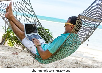 Man In A Hammock On A Tropical Beach Working On Laptop And Talking On The Phone
