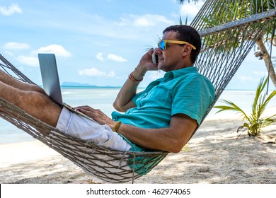 Man In A Hammock On A Tropical Beach Working On Laptop And Talking On The Phone
