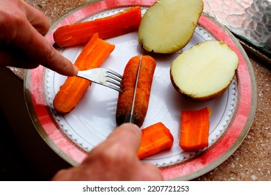 Man hand cuts with knife and fork the cooked potatoes and carrot vegetables in the plate on the kitchen counter preparing homemade food at home with window background - Shutterstock ID 2207721253