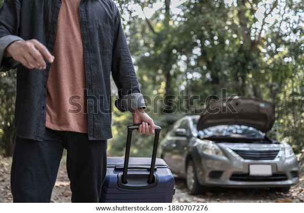 Man\' hand\
drag to travel bag in front of open hood of a broken car on the\
road in the forest. Car breakdown\
concept.