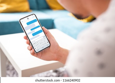 Man Hand Holding The Black Smartphone With Sms App Displayed On A Screen - Modern Frame Less Design In Home Interior, Living Room - Isolated On White Background Angled Position