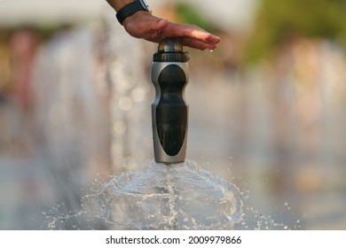 Man Hand Holding A Bottle Of Drinking Water Over The Fountain Jet. It Cools The Water. Hot Summer Day In The Big City. Concept Of The Freshness. Close Up Photography