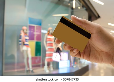 Man Hand Holding Empty Golden Credit Or Debit Card, Member Card, Vip Card With Blurred Background Of Lady Fashion Shop In Department Store, Credit Card Payment Concept.