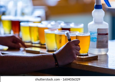 Man Hand Holding A Pen Next Flight Of Craft Four Of Different Beers Glasses On Wooden Trays And Chemical Bottle Close Up, Beer Tasting Room Background