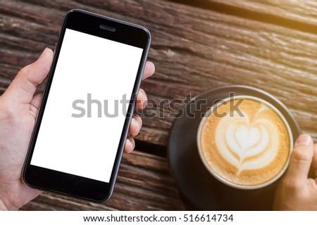 A man hand holding smart phone with latte hot, coffee cup on wood desk in coffee shop. Smartphone with blank screen and can be add your texts or others on smart phone.