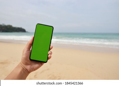 Man Hand Holding The Smartphone With Green Screen Isolated On The Beach Background.