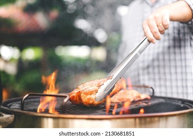 Man hand holding tongs grilling barbecue on fire at backyard on day. Family dinner outdoor style bbq activity.