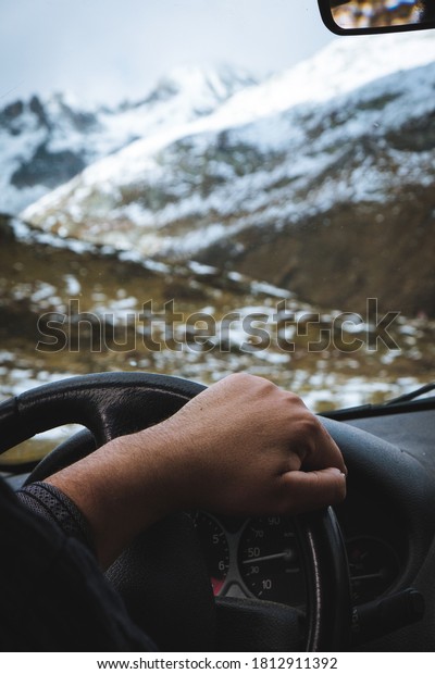 Man with hand on the\
steering wheel as he drives through the snowy, mountainous\
landscape of Switzerland