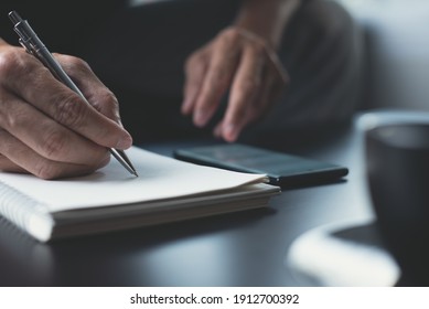 Man Hand With A Pen Writing On Paper Notebook And Surfing Internet On Mobile Phone, Online Working On Table In Office With Dark Background. Business Man Planning Work Project On Notepad, Close Up