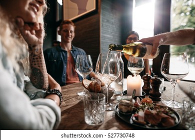 Man Hand Pouring White Wine From The Bottle Into Glasses With Friends Sitting Around The Table. Group Of Young People Having Food And Drinks At Restaurant.